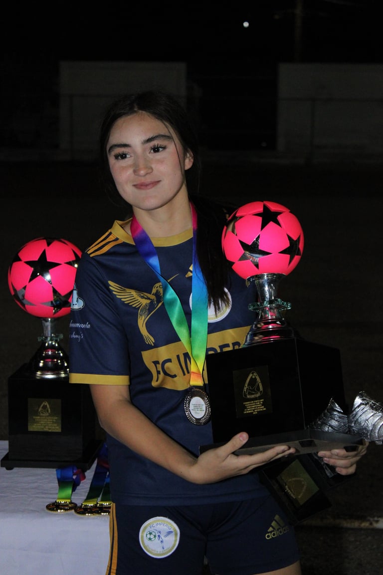 Young female soccer player holding two pink and black soccer ball trophies, wearing navy sports jersey and medals around neck