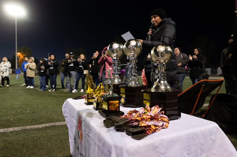Trophy display on white table at nighttime outdoor event with crowd gathered on grass field