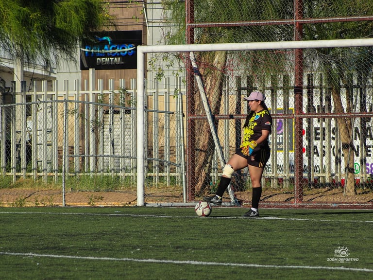 Soccer player in yellow and black jersey controls ball near goal on grass field with white fence and trees