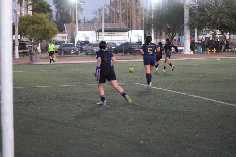Young soccer players in dark uniforms playing on a grass field during daytime with spectators and buildings visible in the background