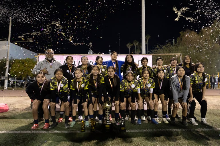 Youth soccer team in black and yellow uniforms posing together at night on a lit field with coaches