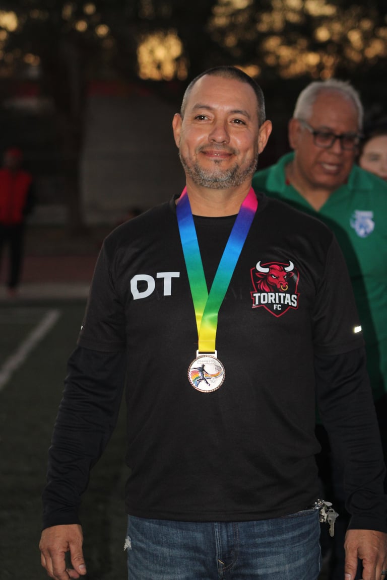 Man wearing black jacket with DT initials and Toritas logo, displaying rainbow medal around neck at outdoor event