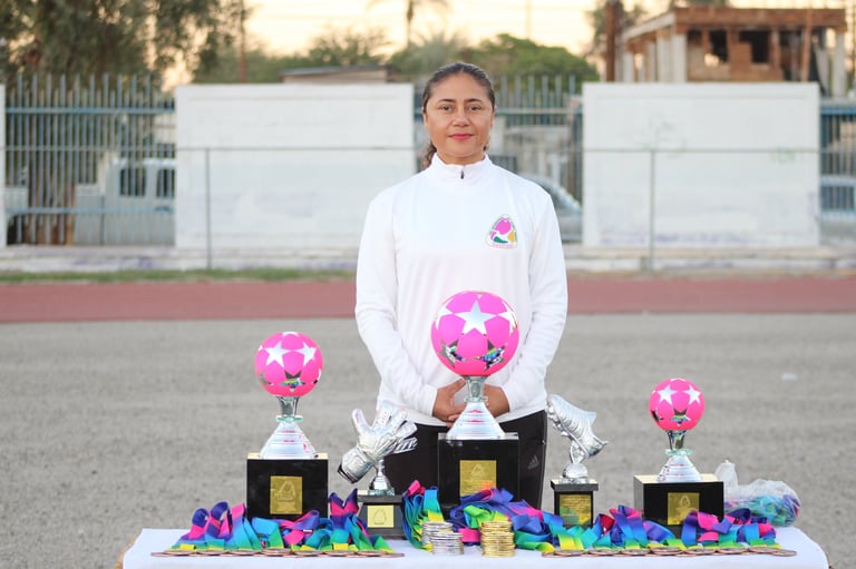 Female athlete holding pink soccer balls with multiple trophies on a table at a sports field