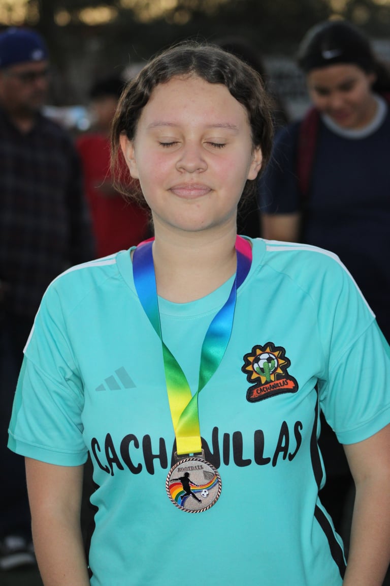 Young athlete wearing turquoise Cachamillas sports jersey with medal around neck, eyes closed, standing outdoors with spectators blurred in background