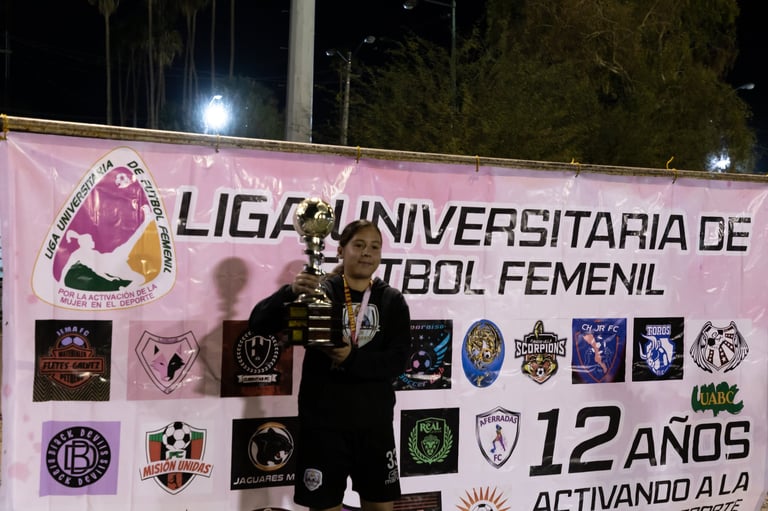 Woman holding trophy at night in front of a pink banner for Liga Universitaria de Futbol Femenil with team logos