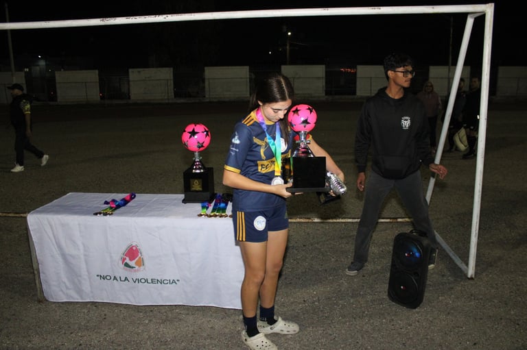 Young soccer player holding trophies at night on a sports field, with teammates and coaches nearby