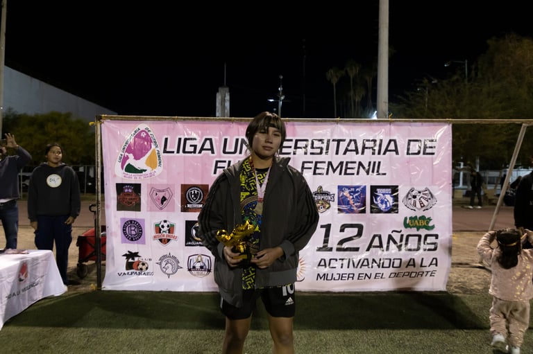 Female athlete holding trophy at night event with banner for womens university soccer league, surrounded by spectators and logos
