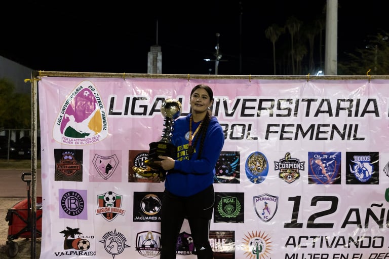 Woman in blue jacket holding trophy standing in front of pink University Womens Soccer League banner with team logos at night