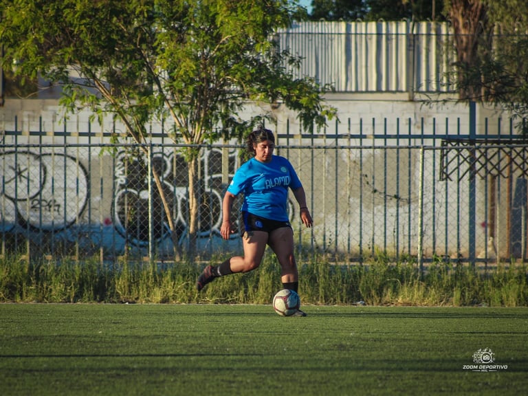 Young soccer player in blue shirt running with ball on grass field with fence and trees in background