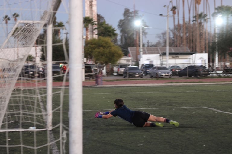 Soccer goalkeeper diving horizontally to make a save on a field with palm trees and parked cars visible in the background