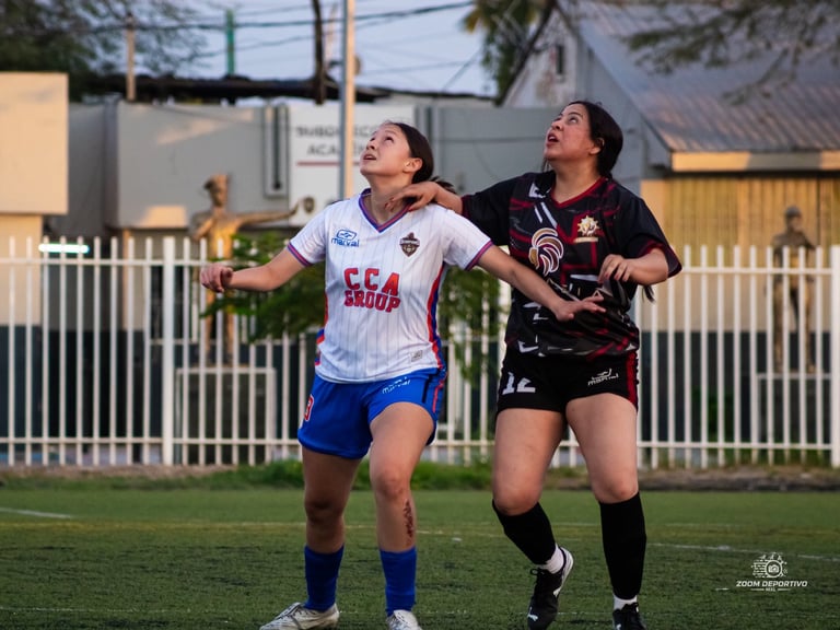 Two female soccer players on a grass field during a match, one in white and blue uniform, one in black, both looking upward