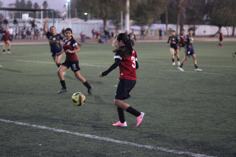 Youth soccer player in red and black jersey controlling ball during match on grass field with teammates and spectators in background