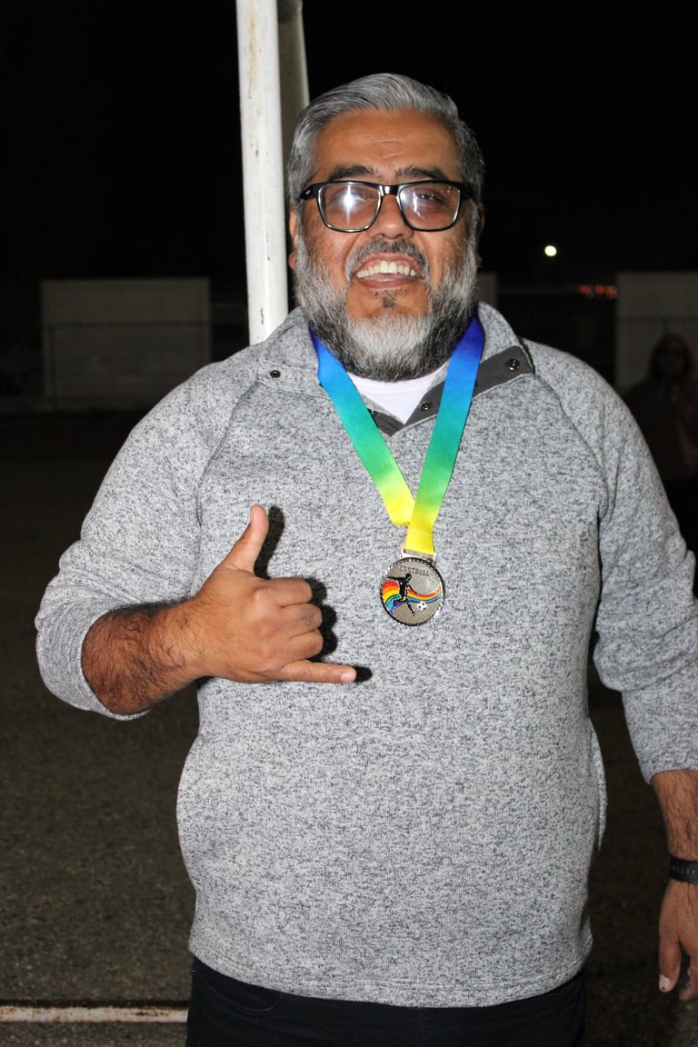 Man with gray beard wearing glasses and gray sweater, holding a medal with rainbow ribbon, giving thumbs up