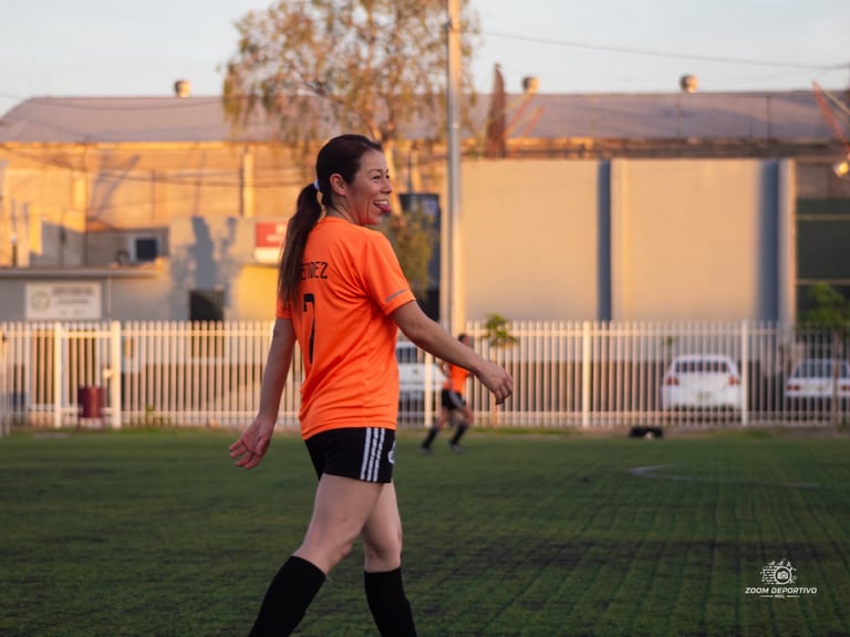Young soccer player in orange jersey running on grass field during sunset with buildings and fence in background