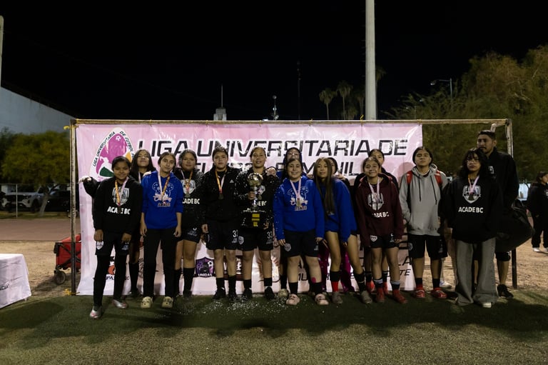 A group of young athletes stands together at night in front of a university sports banner, holding trophies and wearing medals