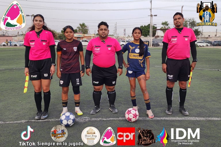 Five female soccer referees in pink and black uniforms standing on a soccer field with sponsor logos displayed below
