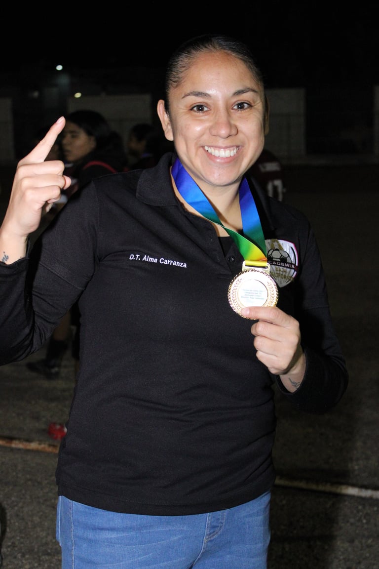 Woman named Alma Carrera wearing a black jacket and jeans, proudly holding a silver medal on a blue and green ribbon, giving a thumbs up gesture