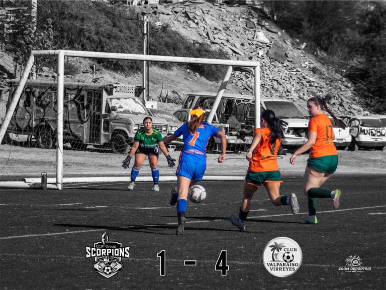 Female soccer players in blue and orange jerseys competing on field with goalkeeper in green, scoreboard showing 1-4, mountains and vehicles in background