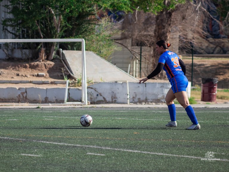 Soccer player in blue jersey controls ball on field near white goal post and bleachers