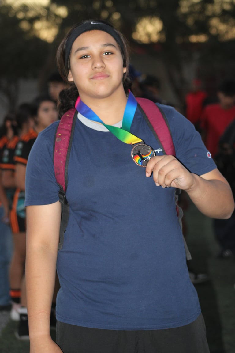 Young person wearing a blue shirt holding a rainbow medal with lanyard around neck at an outdoor event