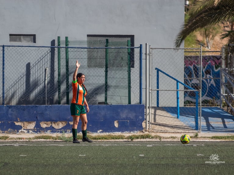 Young soccer player in orange and black uniform stands on field with ball near blue and green fenced facility