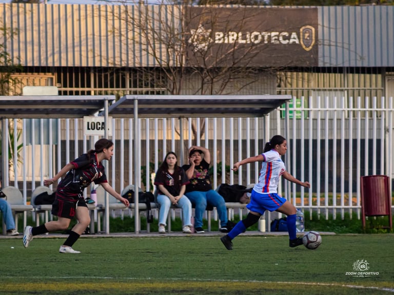 Female soccer player in blue kicking ball while opponent in black defends during outdoor match near biblioteca building