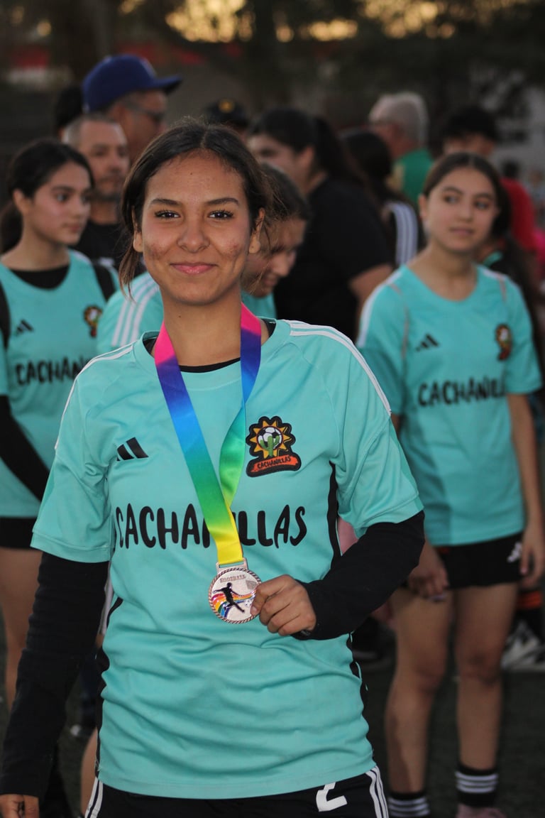 Young athlete in turquoise Cachafillas team jersey wearing a blue and rainbow medal, smiling at camera with teammates in background