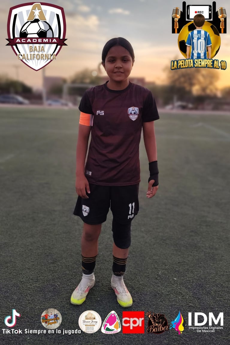 Young female soccer player in maroon and black uniform standing on field at sunset with academy logos