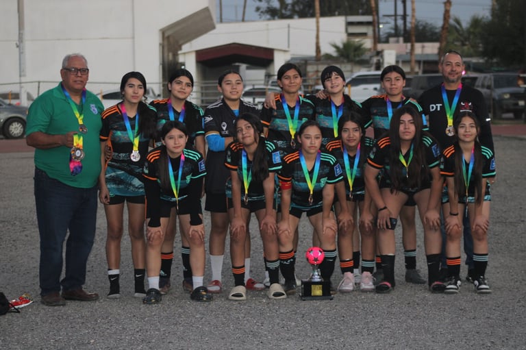 Youth soccer team posing together with coach, all wearing black jerseys and gold medals at outdoor sports facility