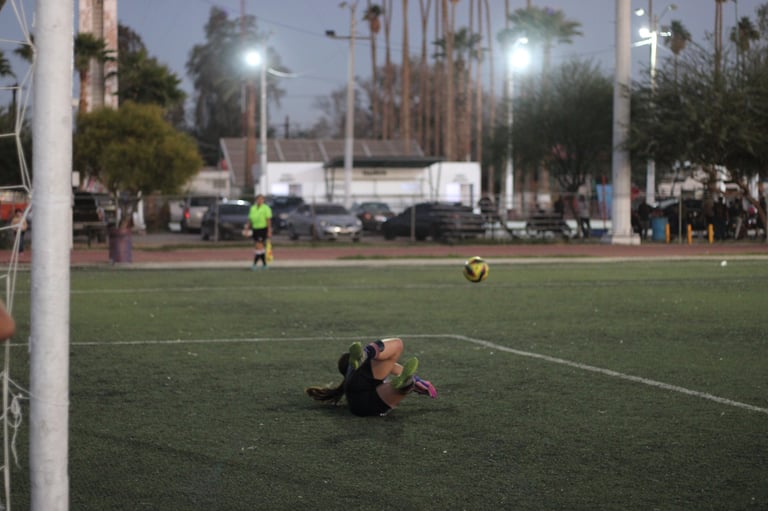 Soccer goalkeeper diving to make a save on a lit field at dusk with palm trees and city buildings in background