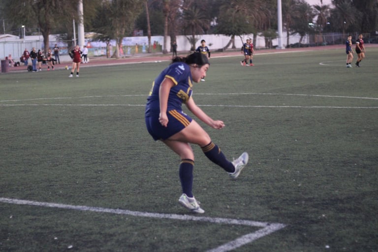 Female soccer player in navy and gold uniform kicking ball on field during match with other players in background