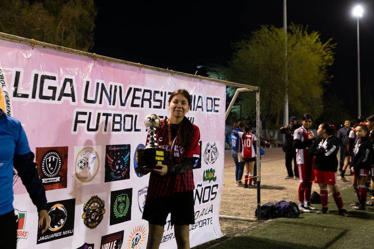 Young athlete in maroon jersey poses in front of Liga Universitaria de Futbol banner at night soccer event