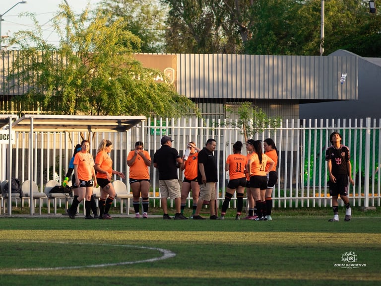 Youth soccer team in orange and black jerseys gathered on sidelines during a match with industrial buildings in background