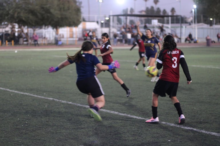 Young female soccer players in dark uniforms competing during a youth soccer match on a lit field at dusk