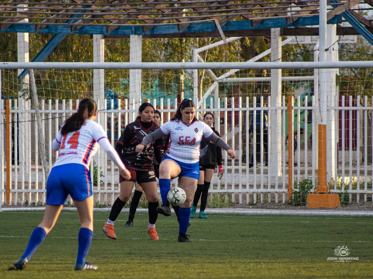 Youth female soccer players competing for the ball during a match on a grass field with white metal fencing in the background