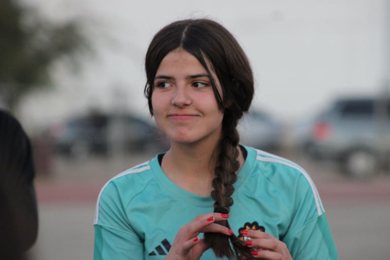 Young person with dark braided hair wearing a turquoise shirt, smiling outdoors in a parking lot